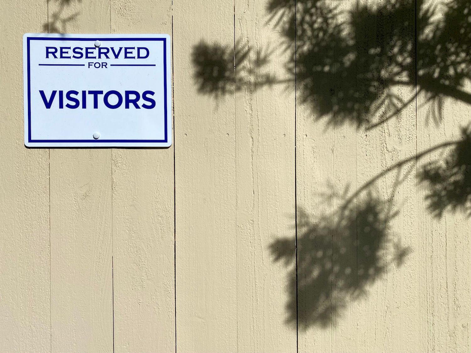 The shadow of a tree on a beige wooden fence, next to a white plastic sign with blue lettering that reads &quot;RESERVED FOR VISITOR&quot;