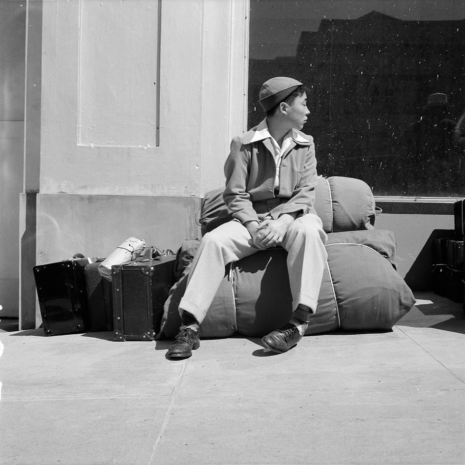 A black-and-white photograph of a teenage Japanese-American boy. He's sitting on duffle bag beside some suitcases, looking out of frame to the right. His body language looks anxious.